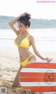 A woman in a yellow bikini sitting on a beach with a surfboard.